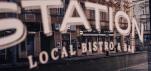 Skiltet «Local Bistro» speiles i et butikkvindu mot en bygate